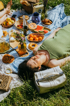 Femme allongée sur l'herbe avec un pique-nique disposé sur une nappe avec fruits d'été, pêches, tomates, herbes fraîches, ambiance soleil du matin